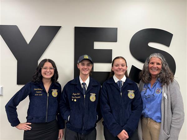  Three students in FFA jackets and a woman in an FFA shirt smile and pose in front of the word YES.